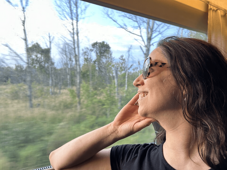 A brunette white woman wearing large sunglasses smiles while looking out of a train window